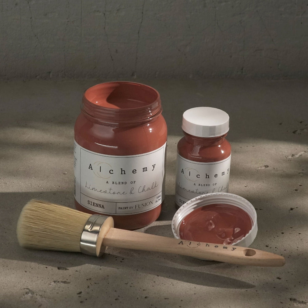 Two jars of rusty Sienna paint from Alchemy by Fusion and a brush on a textured surface