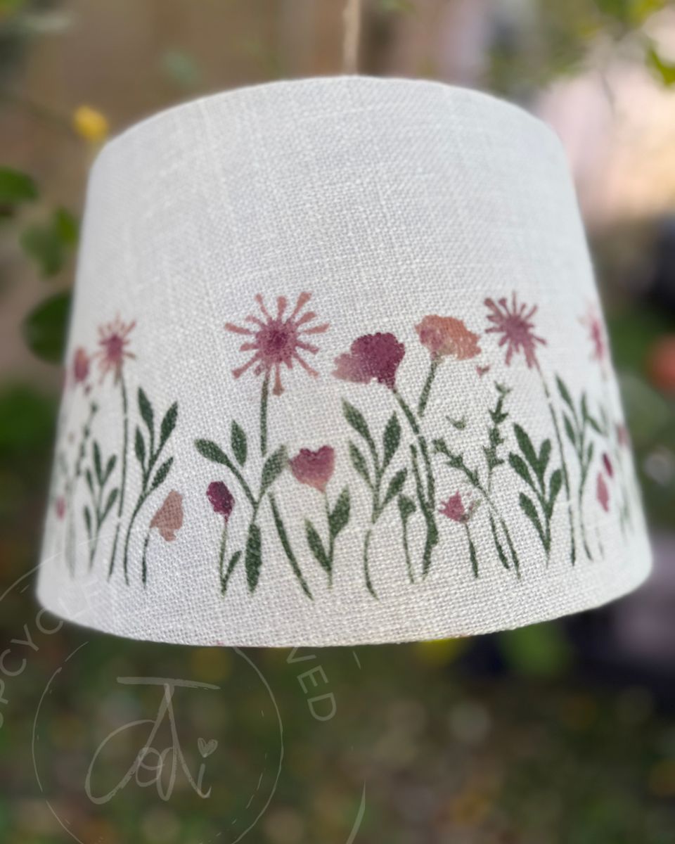 A white fabric lampshade that has been upcycled featuring a painted border of wildflowers in pink, purple, and green, shown hanging outdoors against a blurred garden background. Pink flower lampshade
