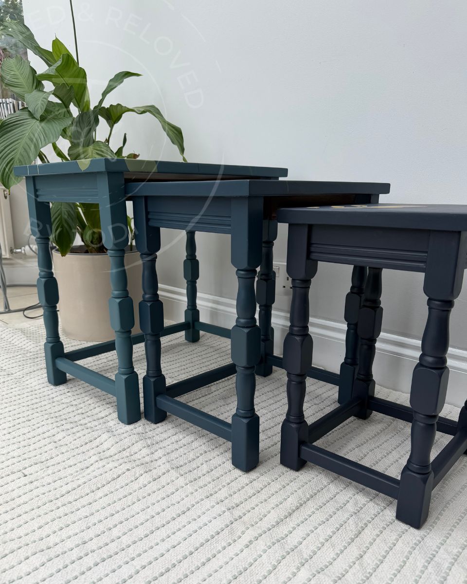 Three nested nesting tables painted in tones of teal to dark blue, styled in a home setting on a pale patterned rug with a potted plant in the background and the watermark logo of Reloved by Jodi a furniture artist in Surrey