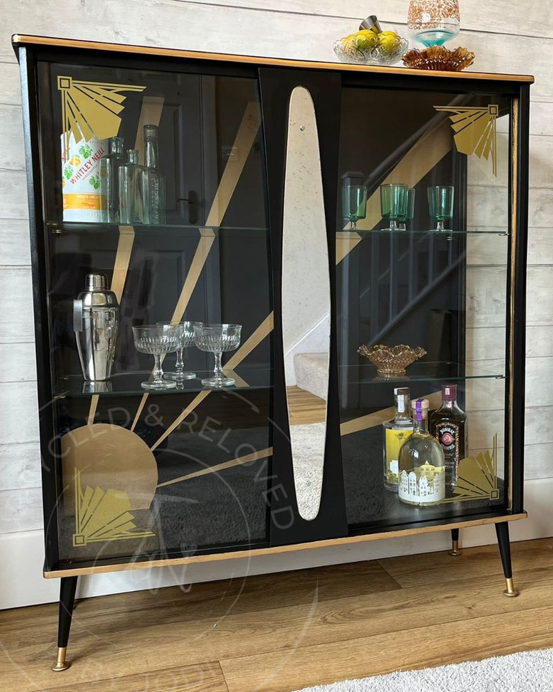 A black and gold Art Deco drinks cabinet with glass doors, glass shelves, gold sunburst detailing and gold-capped legs, transformed by Upcycled & Reloved by Jodi.