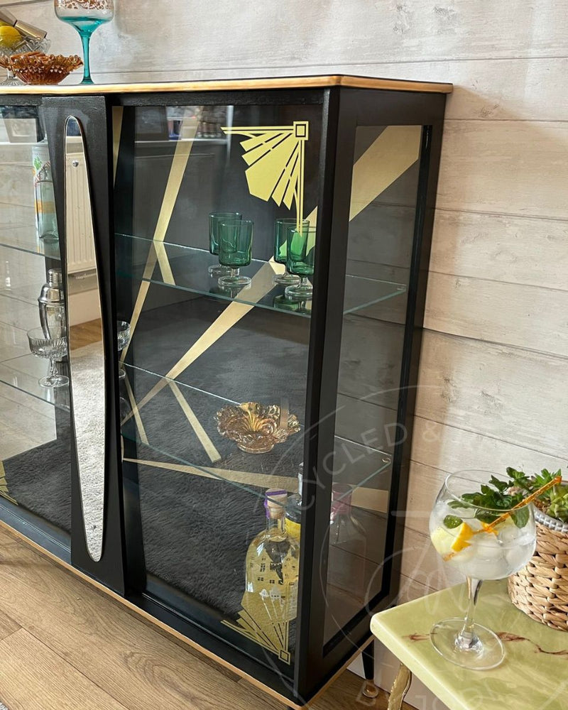 A black and gold upcycled Art Deco drinks cabinet styled with cocktail glasses, bottles, lemons and vintage glassware on a wooden floor, refinished by Upcycled & Reloved by Jodi in Surrey, UK.