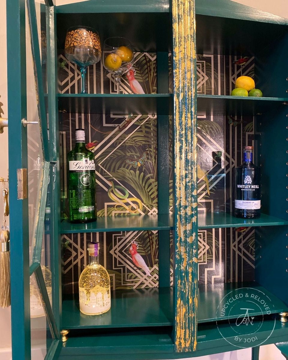 Gin cabinet upcycled in an art deco style, painted in green with gold decorative detailing with gin bottles placed on painted shelves against a wallpapered backing. 