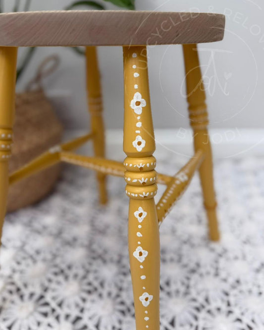 Close up photo of an upcycled stool with a raw pine top and legs painted in Fusion Mineral Paint in Prairie Sunset, a mustard yellow, finished with hand-painted folk-inspired decorative patterns in white. 