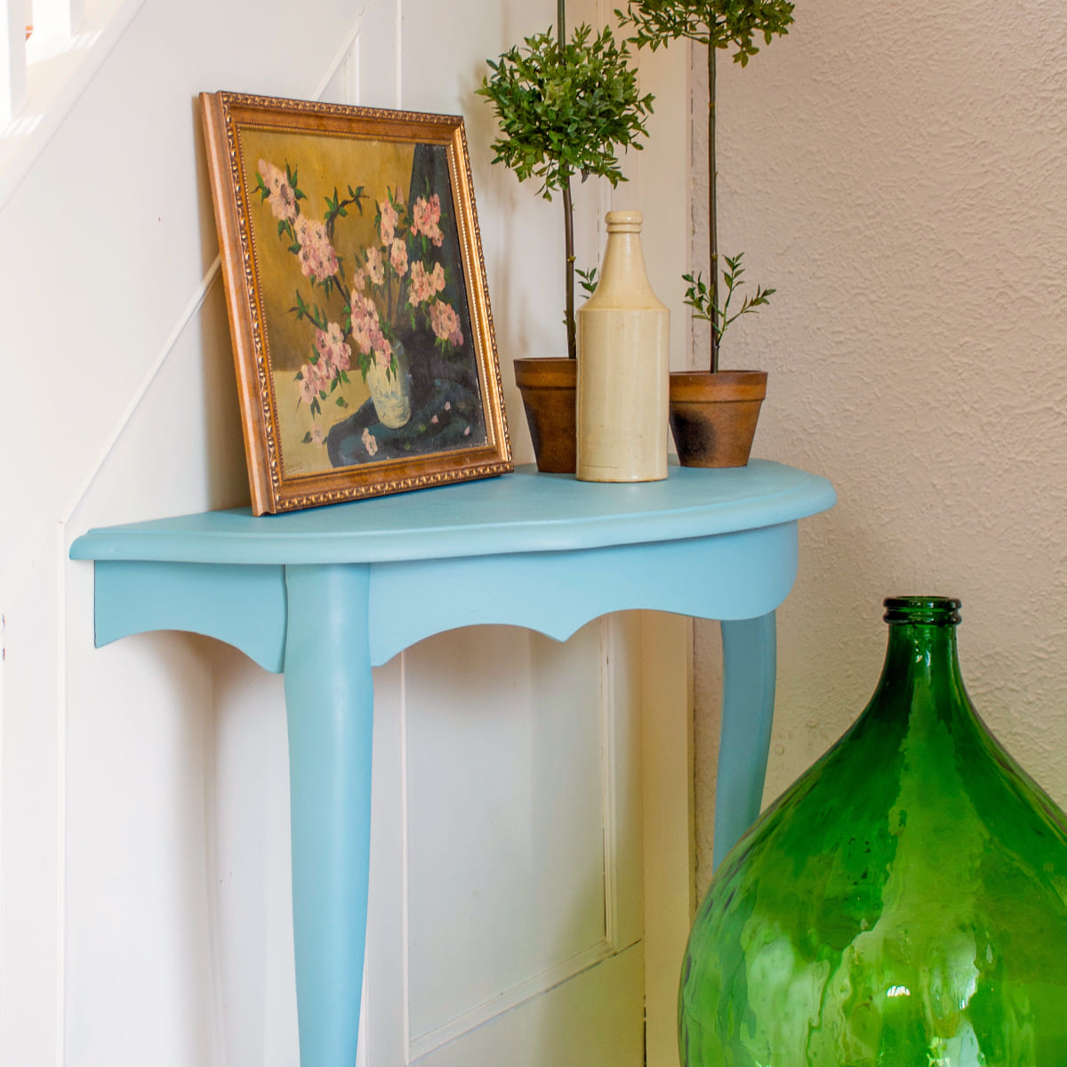 Small table painted in misty Heirloom Fusion Mineral Paint with a framed picture, vase, and green bottle against a white wall.