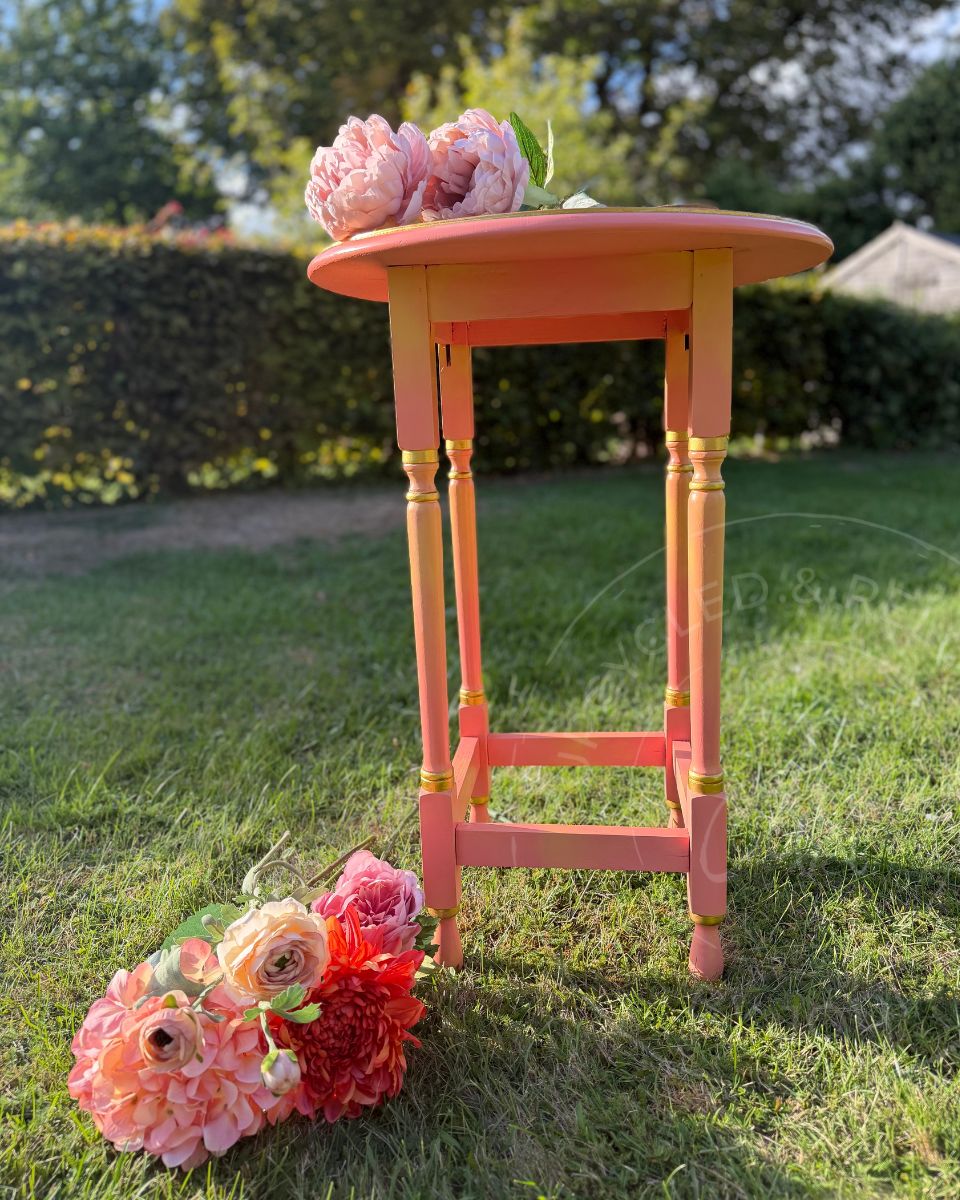 A colourful upcycled side table painted in coral pinks and yellows in a ombre blended effect. the table has a round top and four legs on a squared base. the legs have a gilded detail