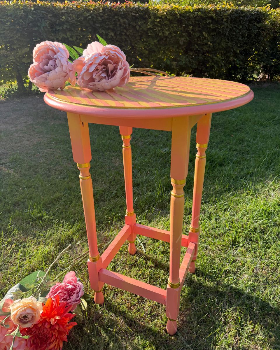 A picture taken in sunlight on a garden of an upcycled round side table painted in coral pinks and yellows. the table has four legs featuring a blended paint effect and gold accents on the trim. A painted piece by Upcycled and Reloved by Jodi in Surrey