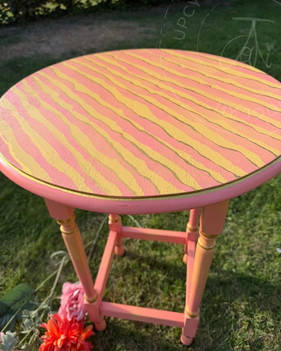 A vintage round occasional table painted in coral pink and yellow tiger or zebra stripes. the pain has a blended or ombre effect using fusion paint