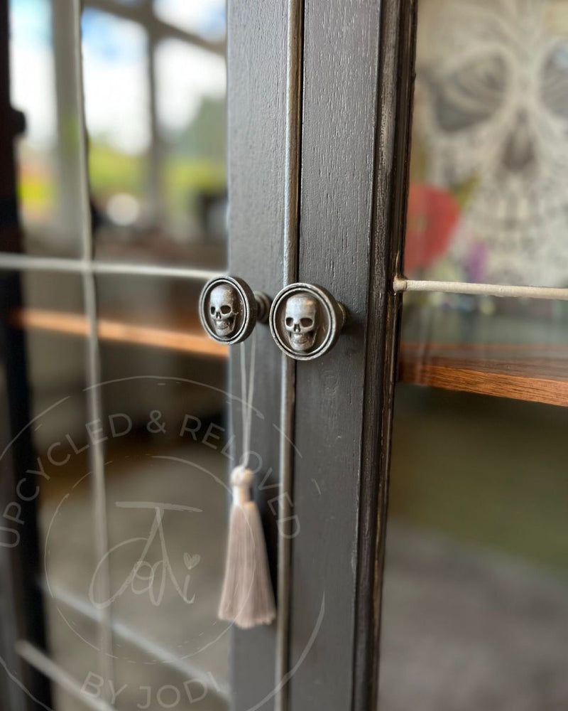 A close-up of skull-shaped cabinet handles on a dark grey painted rum cabinet with silver detailing and glass doors, from Upcycled & Reloved by Jodi in Surrey, UK.