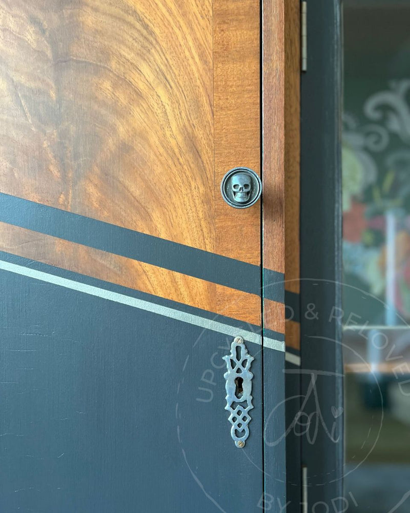A dark grey and wood drinks cabinet with silver geometric lines, skull handles and original glass doors after being upcycled in Surrey, UK by Upcycled & Reloved by Jodi.
