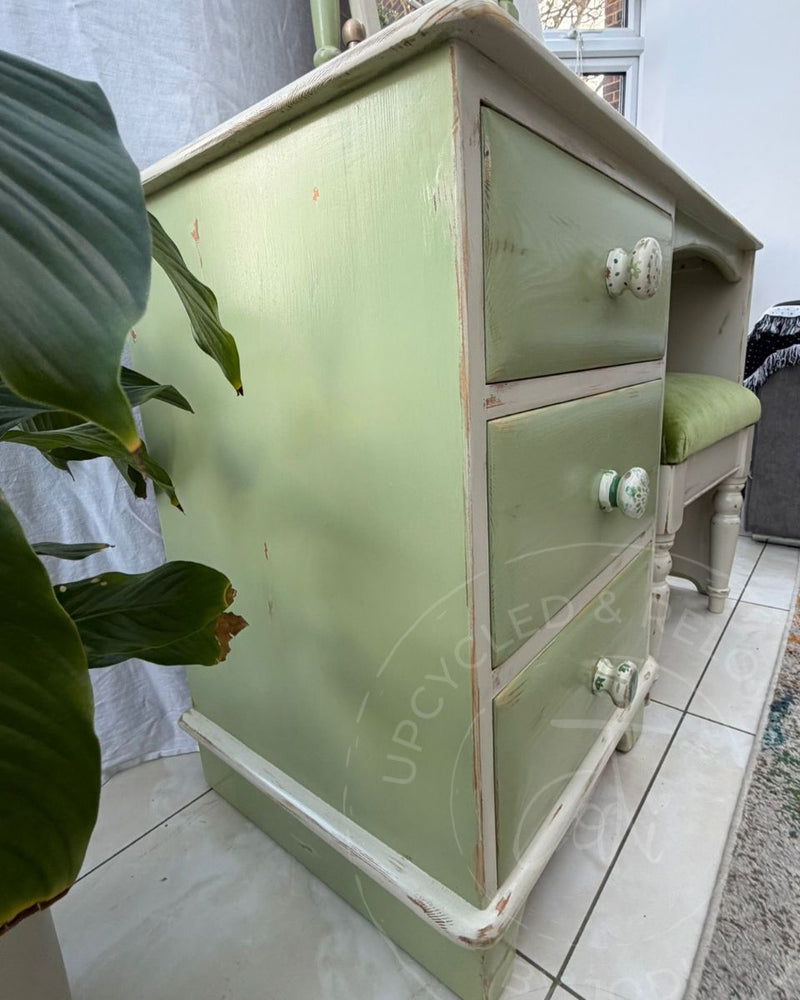 A shabby-chic sage green three-drawer dressing table with quirky ceramic knobs, refinished by Upcycled & Reloved by Jodi, shown in a home setting with a tiled floor, a matching stool, and a large green plant in the foreground. 