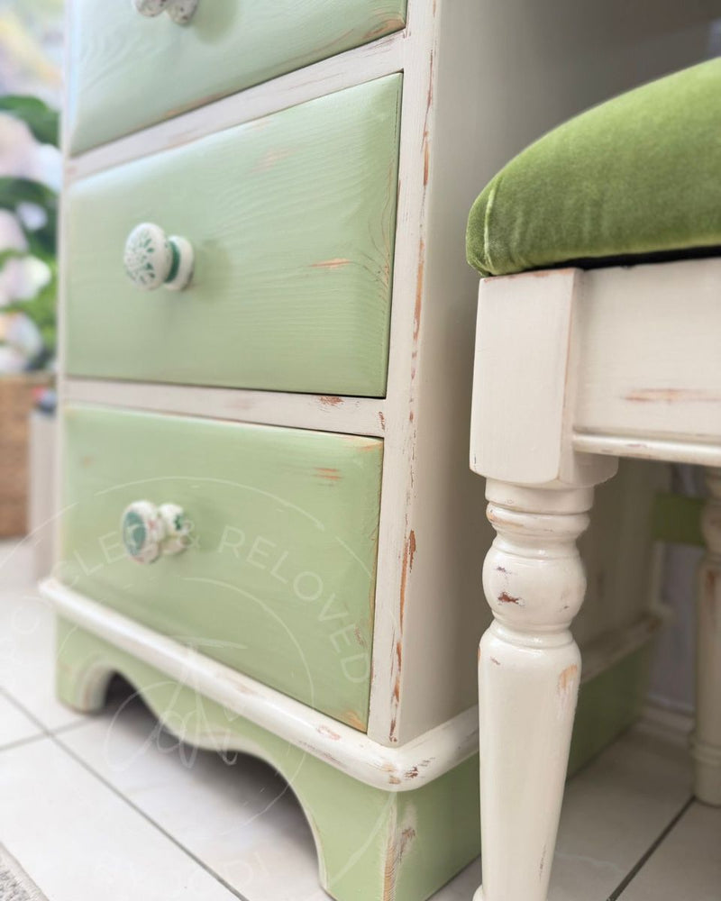 A close-up of a distressed three-drawer dressing table in Fusion Mineral Paint's Upper Canada Green and Champlain cream with a matching stool with a green velvet cushion, refinished by Upcycled & Reloved by Jodi. 