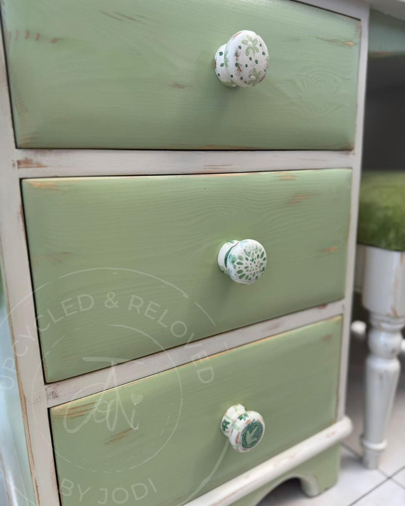 A close-up of a three-drawer dressing table with a distressed sage green finish from Fusion Mineral Paint and white ceramic knobs featuring green floral patterns, restored by Upcycled & Reloved by Jodi.