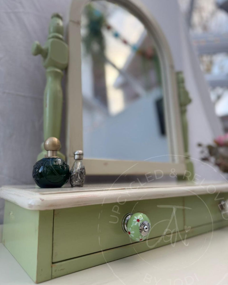 A close-up of an upcycled vanity mirror with drawers, painted in Fusion Mineral Paint Upper Canada Green, a soft sage, and decorative green and white floral ceramic knob. 
