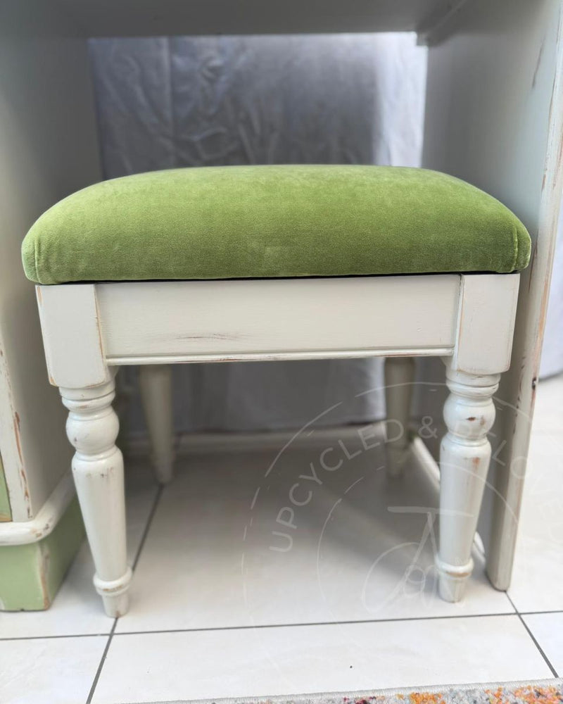 A vintage-style white wooden stool painted in Fusion Mineral Paint's Champlain cream with a distressed finish and a green velvet cushioned seat. 