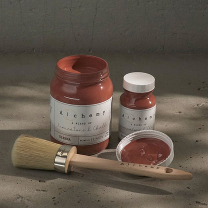 Two jars of rusty Sienna paint from Alchemy by Fusion and a brush on a textured surface