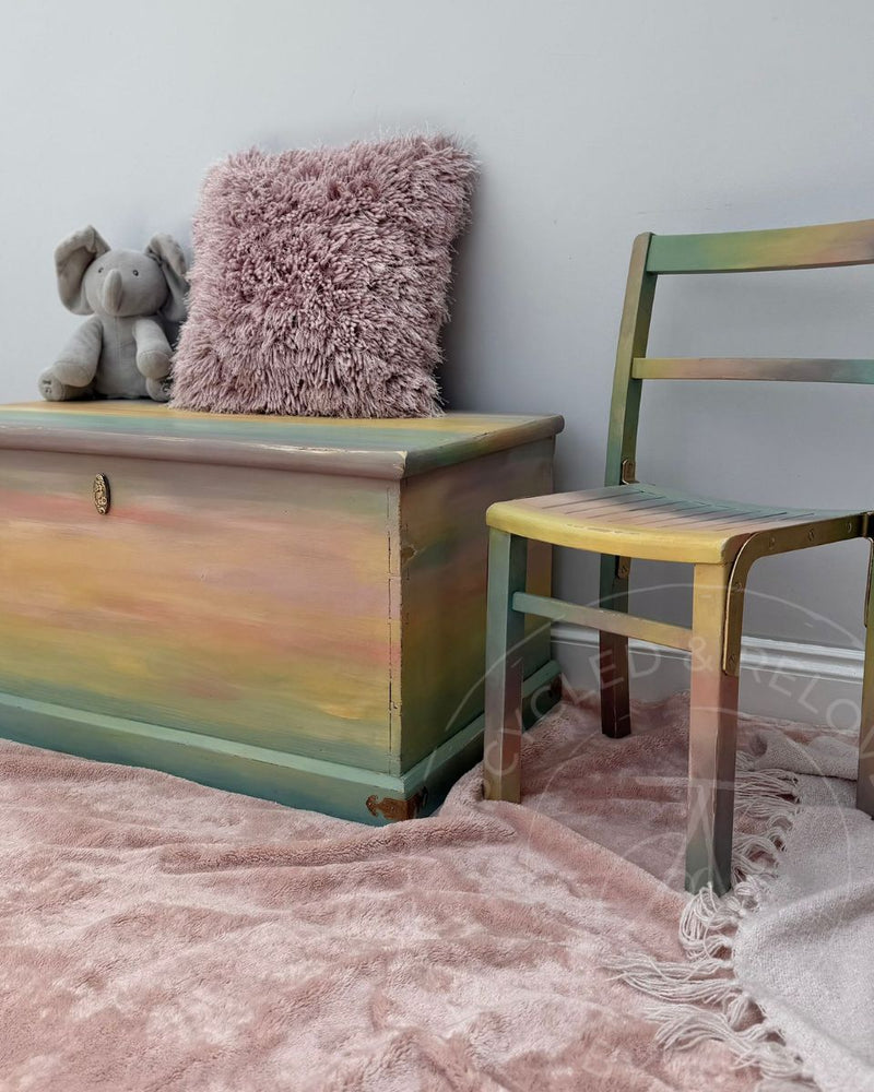 Upcycled children’s toy chest and chair painted in an ombre effect in soft pastel Fusion Mineral Paint shades with a soft toy elephant and pink fluffy pillow atop the chest. 