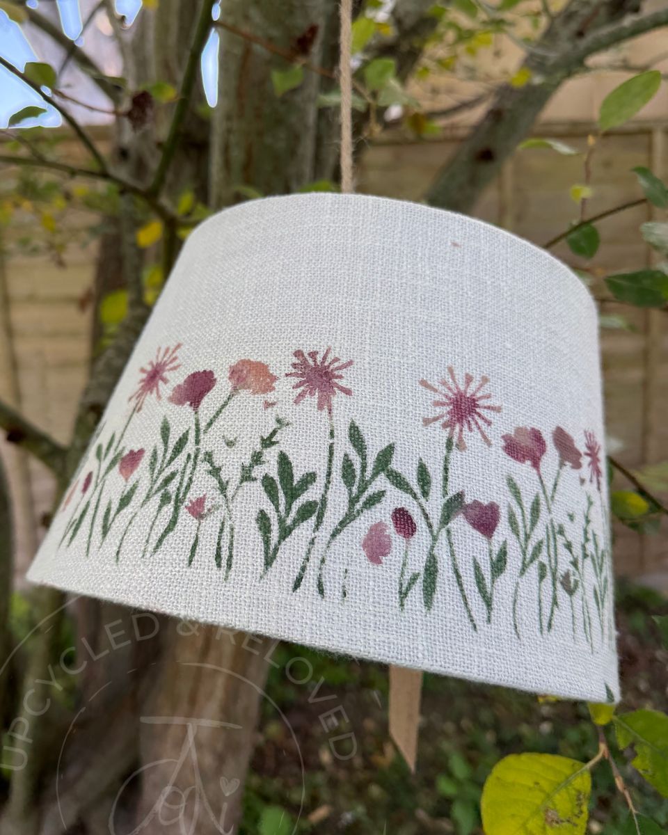 An off-white painted upcycled lampshade with  pink and purple flowers with green leaves on a white background. The logo Upcycled & reloved by Jodi is visible in the bottom corner