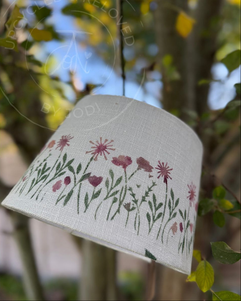 A fabric lampshade with a floral pattern of pink and purple flowers and green leaves, hanging outdoors against a blurred background of trees and foliage. The lampshade is upcycled and reloved by Jodi