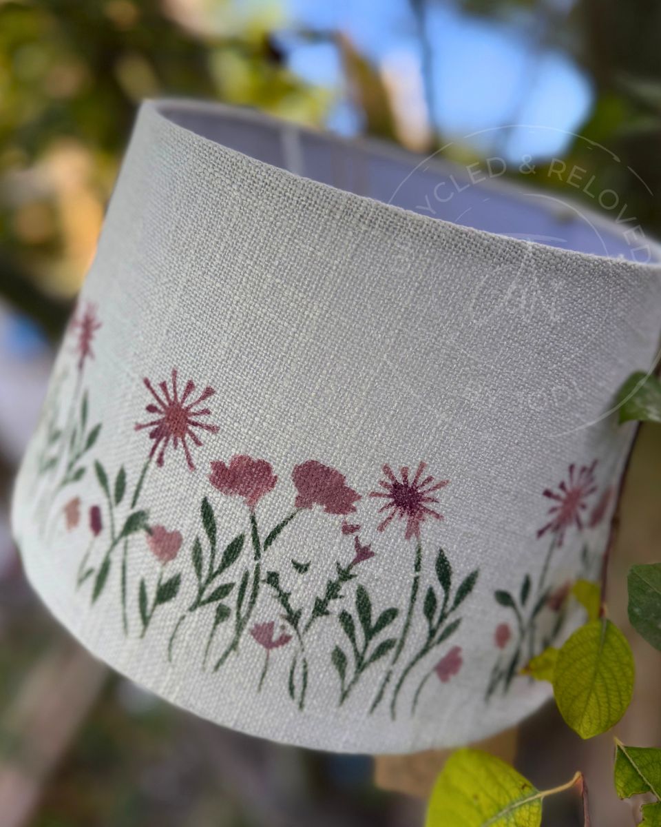 A close-up of a cylindrical lampshade made of textured off-white fabric, decorated with a hand-painted style floral pattern of pink flowers and green leaves, set against a blurred outdoor background of green foliage and blue sky.