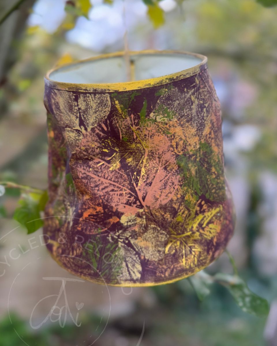 A quirky upcycled lampshade with a leaf print pattern in autumn colours, hanging outdoors against a blurred background of green leaves. The lampshade is a rich burgundy with autumnal colours of leaves and a gold edging