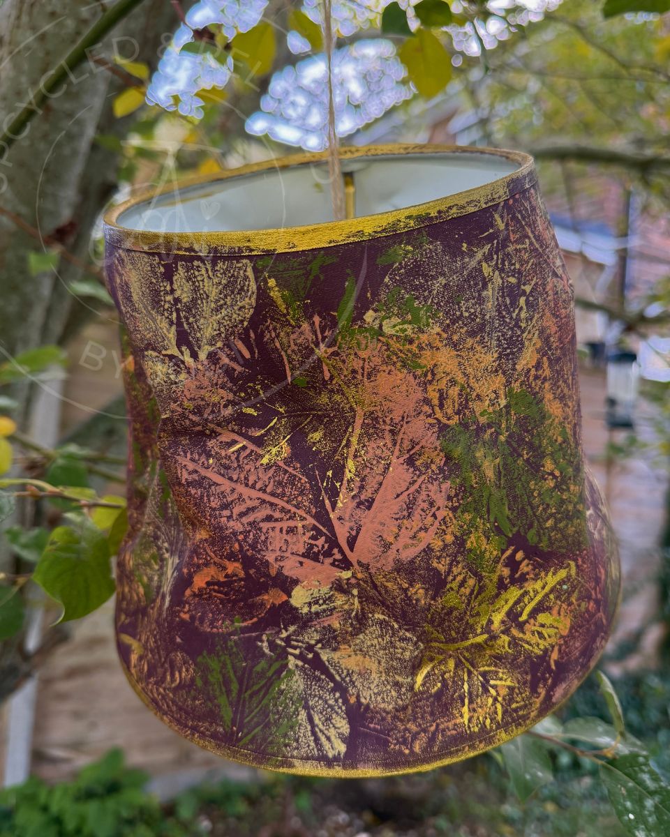 A n upcycled lampshade in burgundy purple with colourful leaf patterns and gold accents. The lampshade has a distressed autumn leaf pattern in purple, coral, green, and gold, photographed outdoors against a blurred garden background.