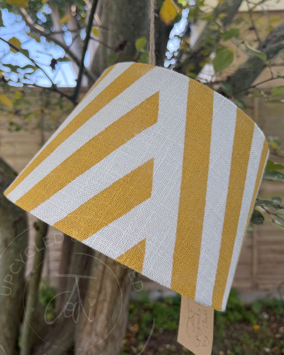 A fabric drum lampshade with a bold yellow and white diagonal striped pattern, hanging outdoors from a tree branch in a garden setting. The lampshade is handpainted by Upcycled and Reloved by Jodi for upcycled home decor