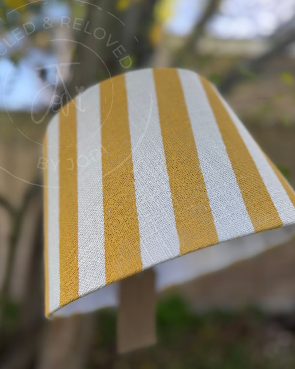 A close-up of a fabric lampshade with vertical mustard yellow and white stripes, photographed outdoors with a blurred natural background. A faint 'Upcycled & Reloved by Jodi' watermark is visible across the image.
