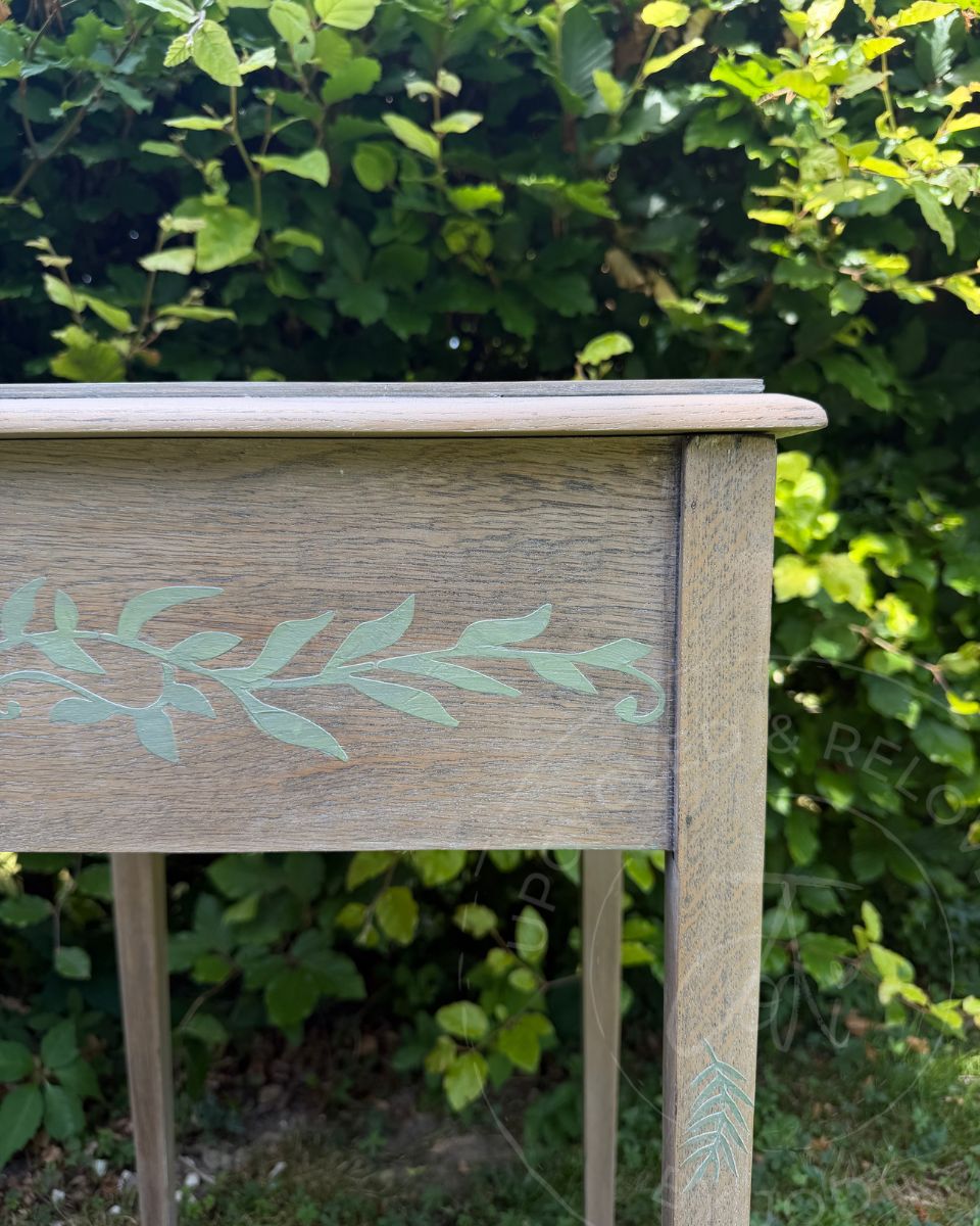 A close-up of an upcycled square oak wood table outdoors, with long square legs, showing a light natural wood grain finish and a raised stencilled sage green leafy branch motif on the front panel, with a lush green garden background.