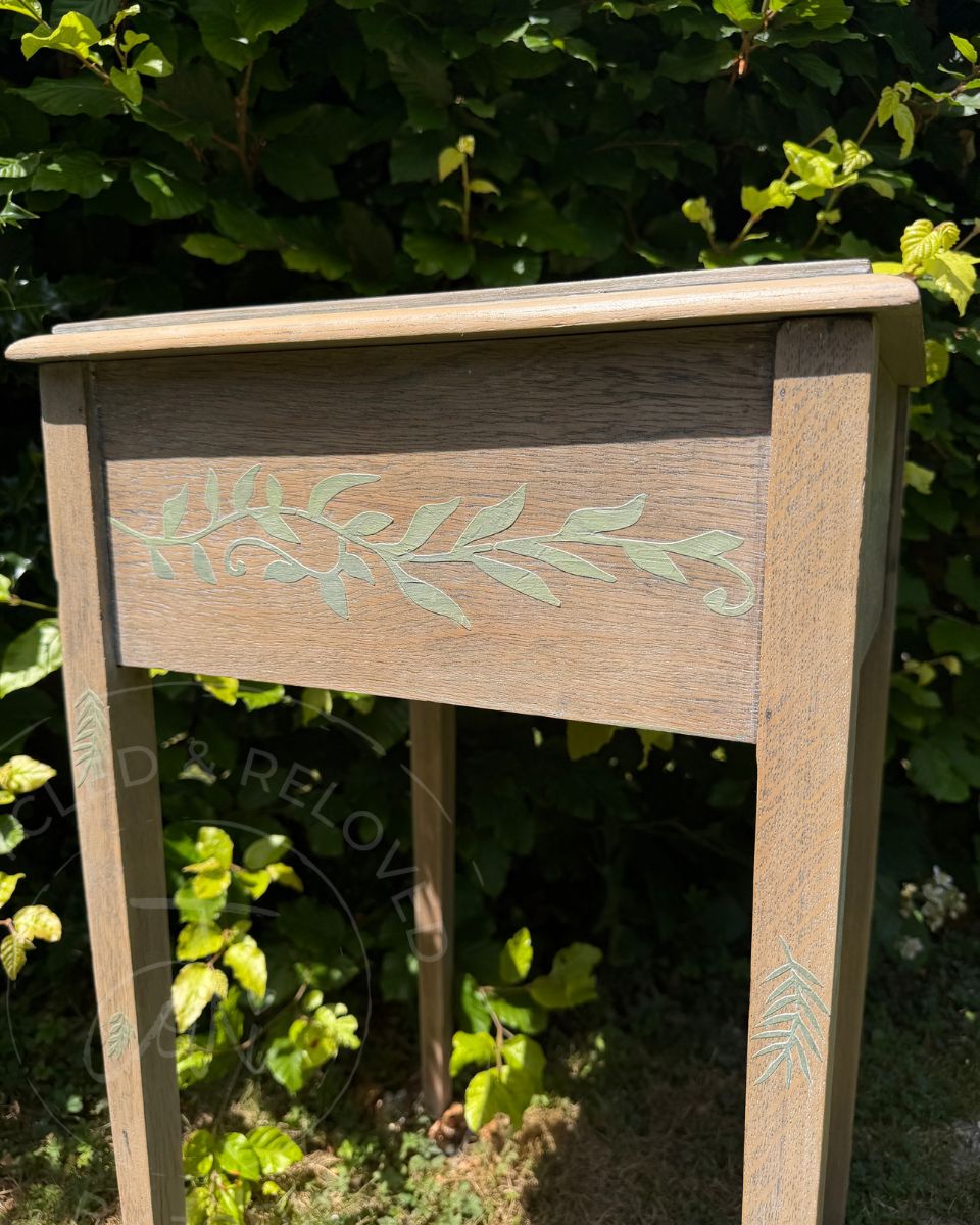 A small wooden upcycled side table or end table with a natural light oak finish and pale green leaf raised design photographed outdoors in front of dense green foliage. The overlapping lid lifts for storage