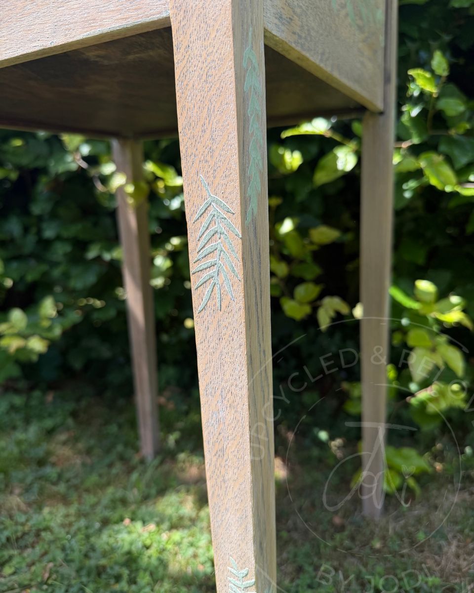 Close-up of a light oak wooden table leg featuring a light green leaf design, set against a blurred background of green foliage and grass.