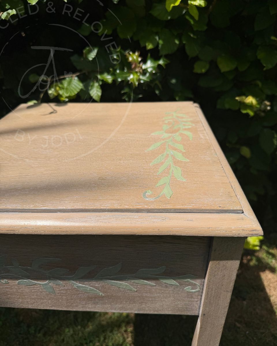 A light oak wood square side table with pale green leafy raised stencil designs, photographed outdoors in front of green foliage. The wood grain shows a whitewash effect