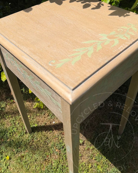 A top view of an upcycled vintage wooden sewing table with a distressed light brown top showing the light wood grain and pale green botanical leaf designs on the top and sides photographed outdoors on grass in direct sunlight. The logo Upcycled and Reloved by Jodi is visible