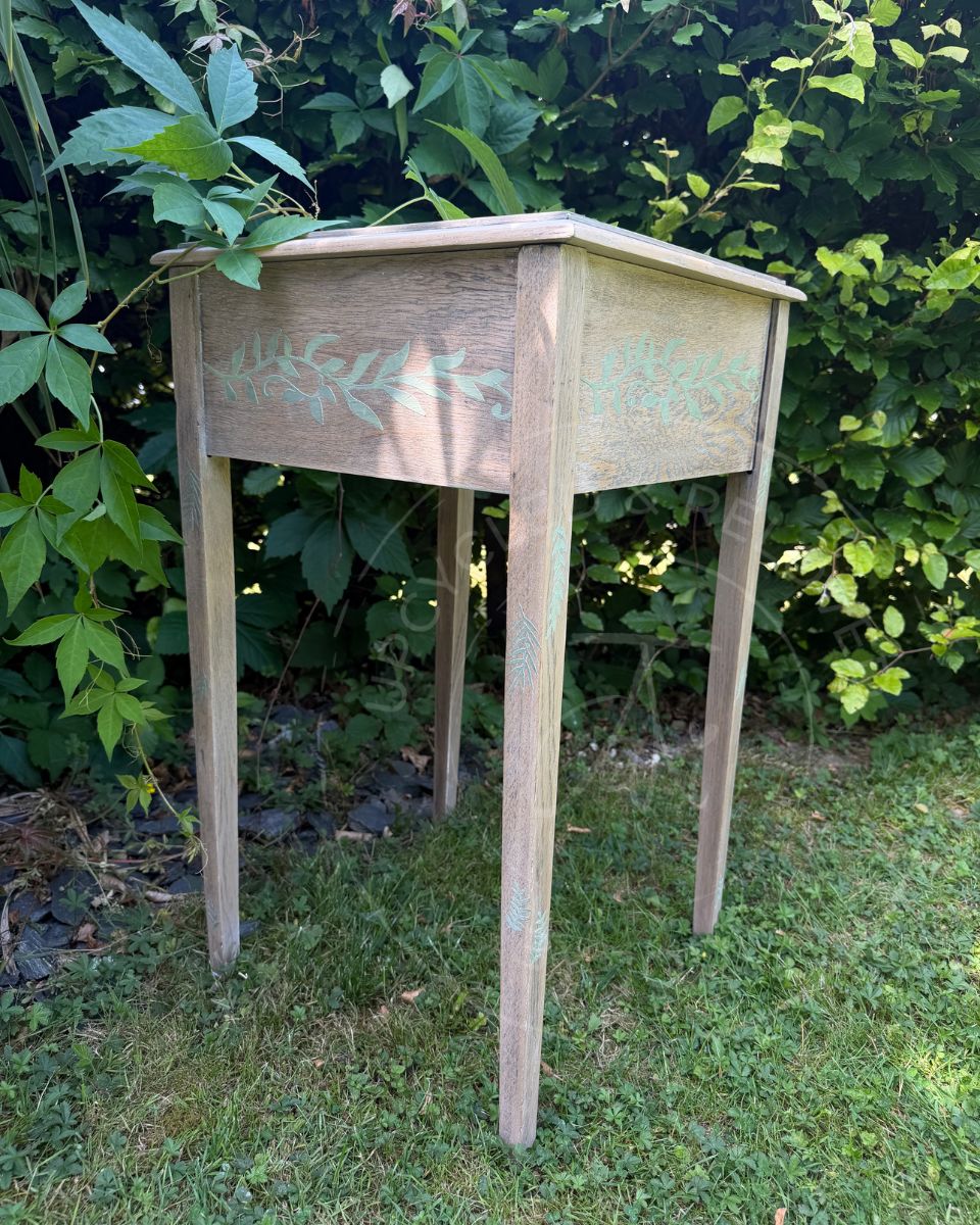 A small light coloured wooden side table with four legs and a hinged top, featuring a pale light washed wood  finish and faded green botanical leaf designs in pale green raised stencil pattern on the side panels, set outdoors on grass with green foliage in the background.