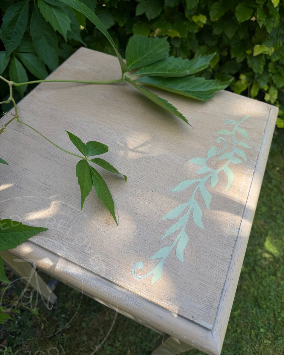 The top of a square wooden sewing box table with a light wood upcycled finish and a mint green leafy vine design on the lid, photographed outdoors on grass with green leaves draped over it.