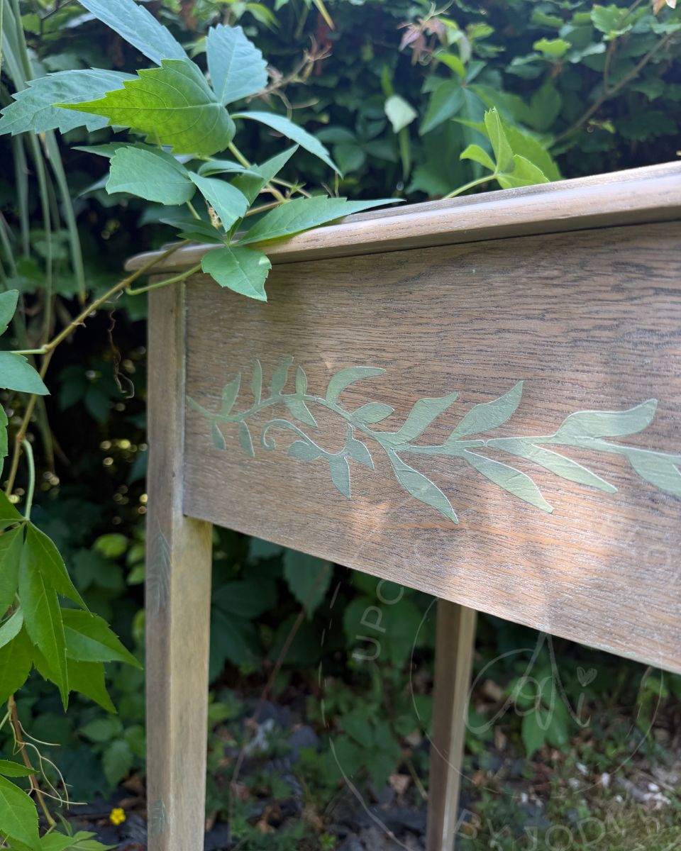 A close-up of a paint-washed wooden upcycled sewing table with a light green leafy branch design, photographed outdoors with green foliage in the background.