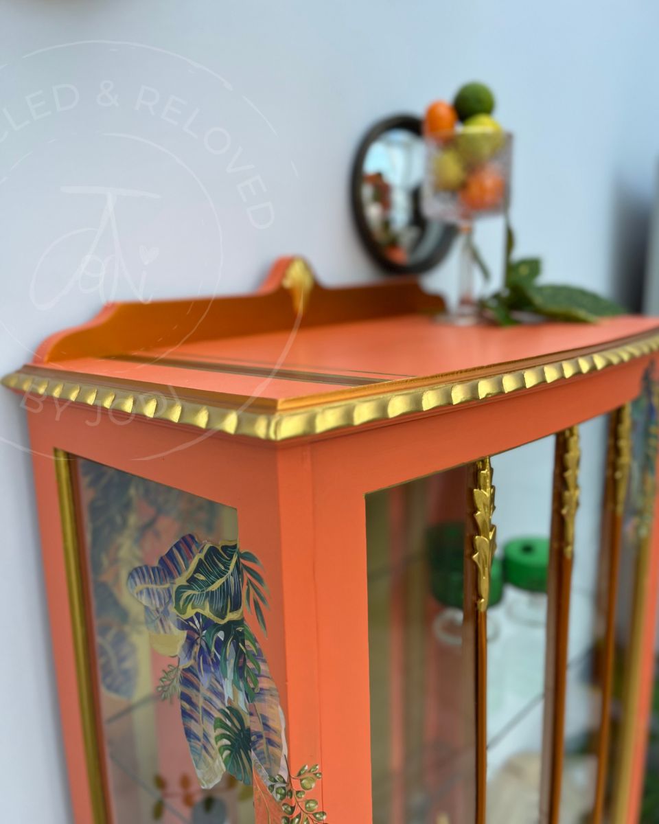A refinished decorative cocktail cabinet or display case with a bright orange body, gold trim, and glass doors featuring tropical leaf illustrations. The upcycled cabinet is shown in an indoor setting with a mirror and fruit on top. The logo 'Upcycled & Reloved by Jodi' is partially visible