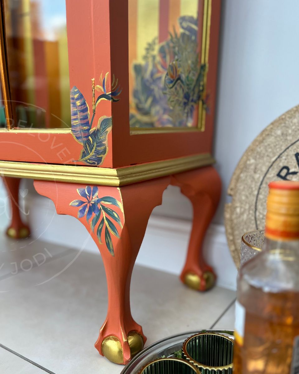 A close-up of an upcycled drinks cabinet or  decorative display case with a bright orange painted body, gold trim, and tropical leaf motifs in blue, green, and gold. The statement piece of furniture is shown indoors on a light floor. It is upcycled by Jodi Walker, a furniture artist in Surrey