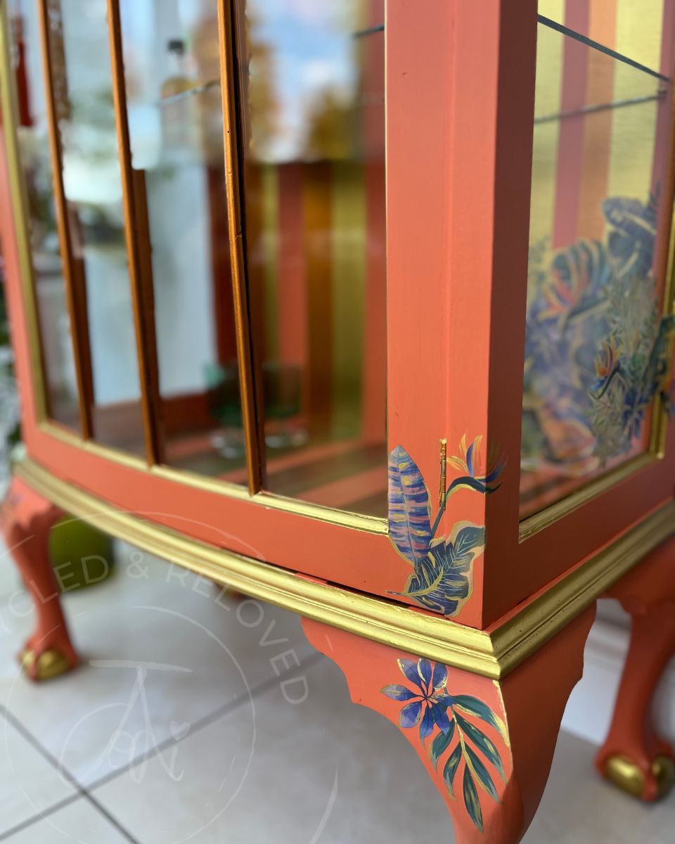 The base and side view of a vibrant drinks cabinet, display cabinet or cocktail cupboard with tuscan orange painted wood, gold trim, and tropical floral motifs on the lower corner supports. The piece of furniture has Queen Anne style legs with ball and claw feet. 