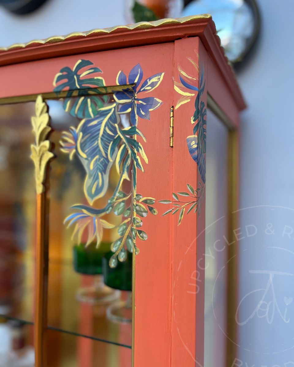 A close-up of a refinished wooden cocktail cabinet in bright orange with gold trim and tropical leaf transfers in blue, green, and gold on the glass door. The statement piece has gold accent on the doors and edges