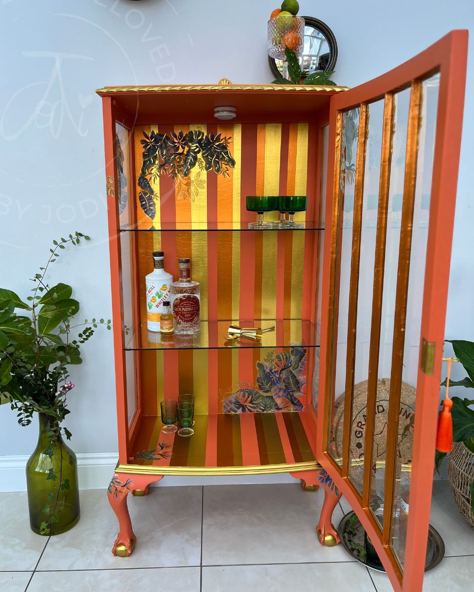 A tall, narrow glass-fronted cocktail cabinet with a bright orange frame and gold vertical stripes, shown open with glass shelves. The cabinet is decorated with floral motifs, the interior has bold metallic stripes in gold and copper and holds a few bottles and glasses. LED lighting is visible inside 