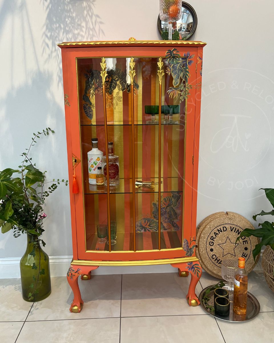 A professionally refinished orange glass-front cocktail cabinet with gold trim, gold and copper stripes and carved legs, styled in a home setting with plants, bottles, and a tray on a tiled floor.