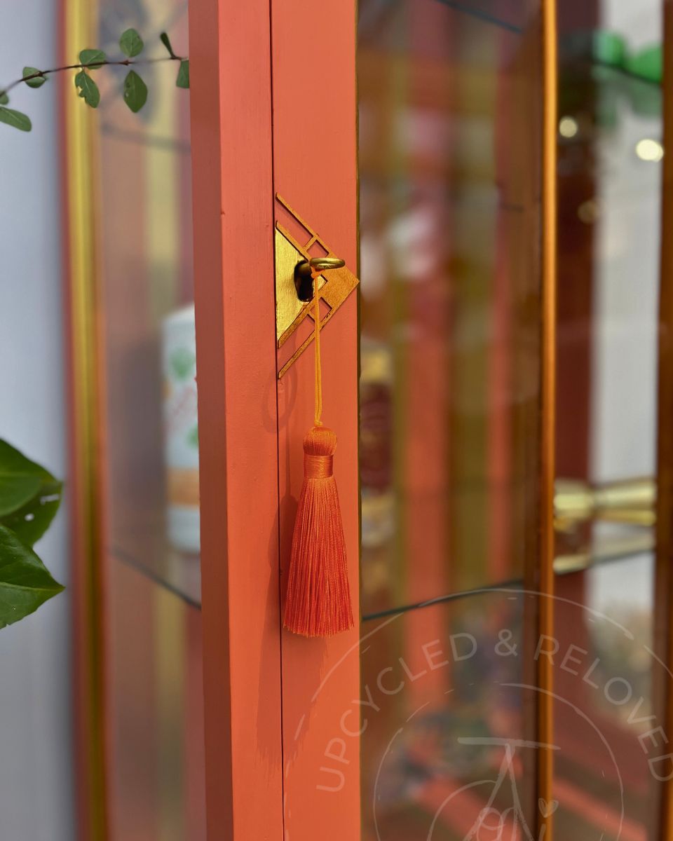 A close-up of a vibrant orange wooden cabinet or display case with a gold geometric accent and a matching orange tassel hanging from the handle. The background shows blurred glass doors and warm interior lighting, with some green leaves visible at the edges.