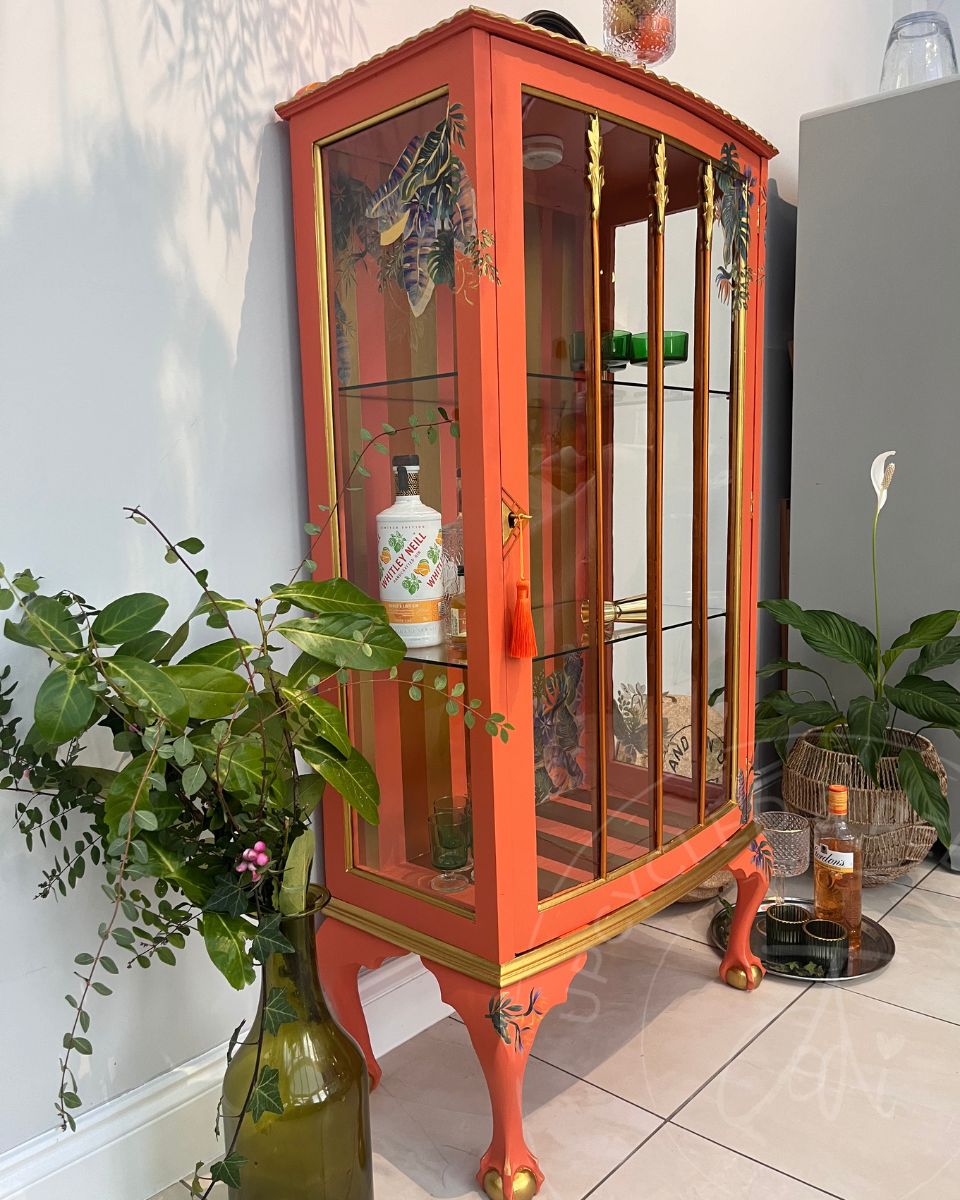 A tall, glass-fronted orange drinks cabinet with gold trim and Queen Anne style carved legs, styled in a home interior next to potted plants and holding alcohol bottles. An example of professionally refinished furniture by Upcycled and Reloved by Jodi