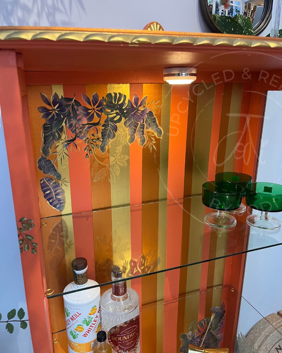 A decorative upcycled orange and gold striped cabinet with tropical leaf motifs on the glass doors. The cabinet holds drinks bottles and glasses, there are copper and gold metallic stripes on the interior and gold accents on the fluted edge.
