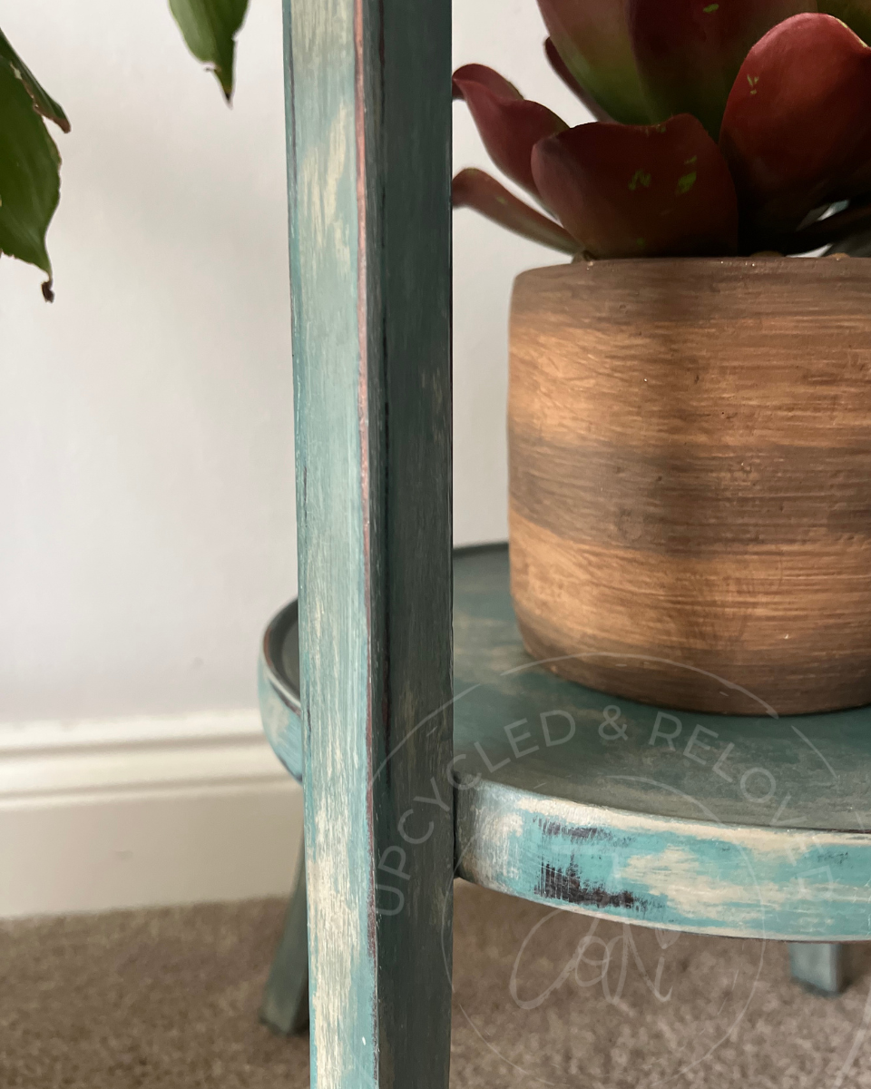 A close-up of a distressed teal blue green wooden side table or plant stand holding a red succulent, set against a white wall and beige carpet. The upcycled plant stand is aged with layered paint.A faint 'UPCYCLED & RELOVED' watermark is visible.