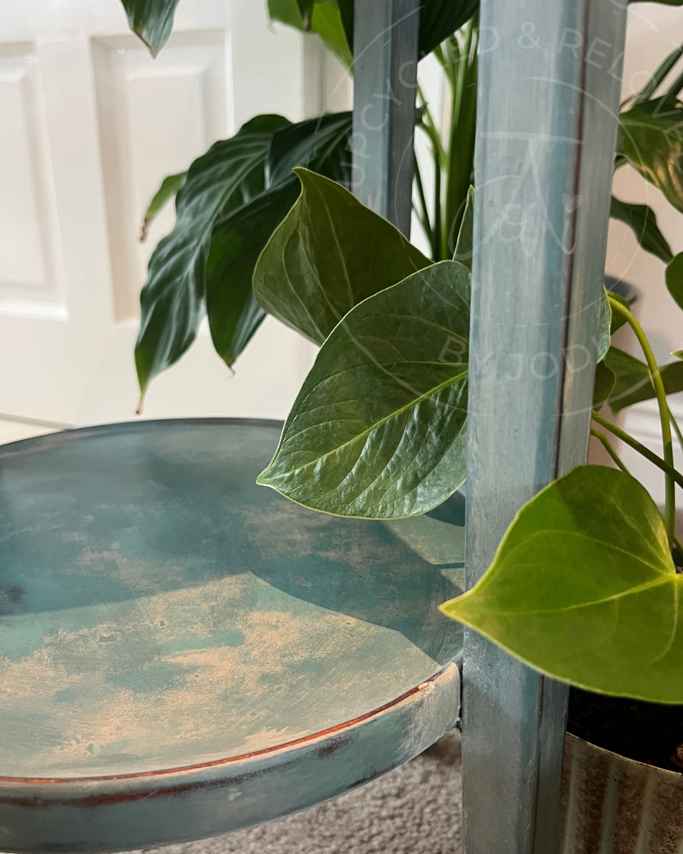 A close-up of a round vintage plant stand in an aged,  distressed teal-blue and green milk paint. The upcycled table has worn edges and a smooth finish in soft sage greens and blues