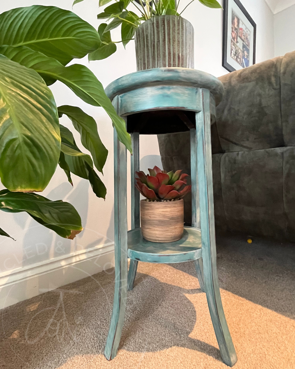 A distressed teal blue wooden two-tier vintage side table or plant stand styled in a living room setting, holding a potted green plant on top and a small succulent on the lower shelf. The shabby chic table has a smooth finish and a naturally aged appearance