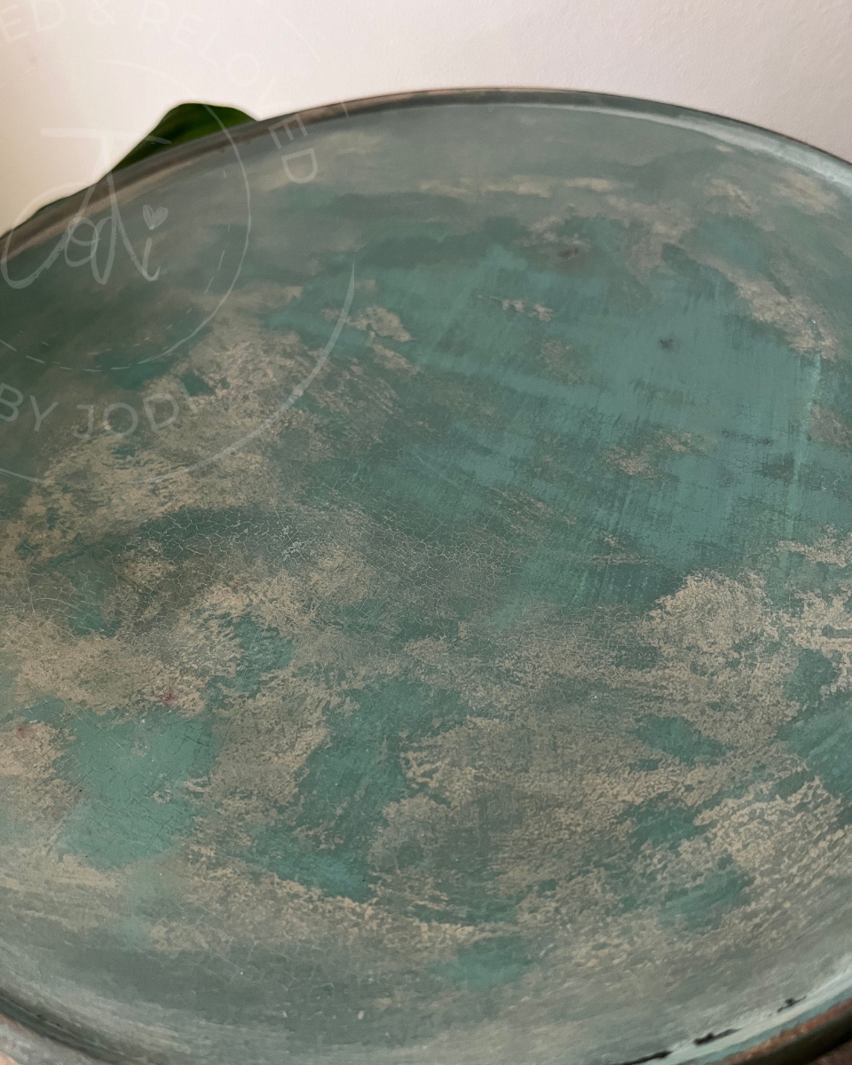 A close-up of a distressed teal-green painted plant stand, showing weathered grey and white wear patterns. An aged vintage plant stand upcycled and reloved by Jodi