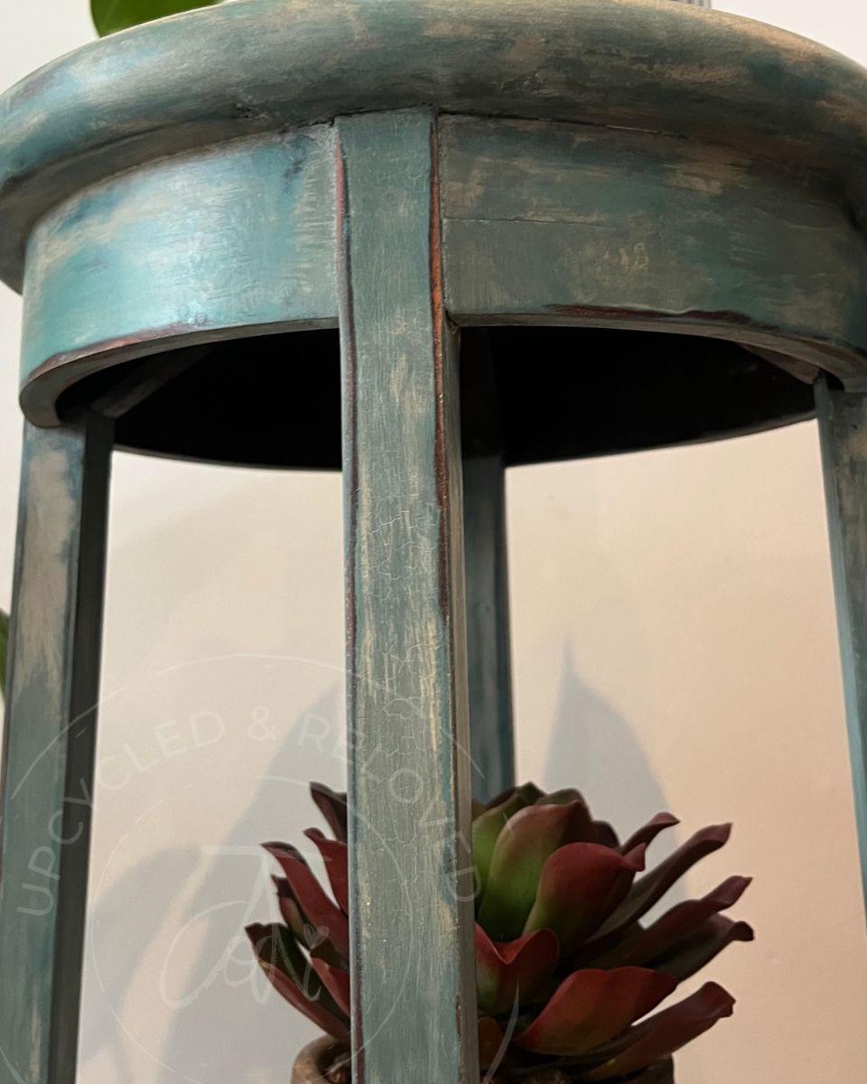 Close-up of a distressed teal-green vintage plant stand with a succulent plant visible inside, with the logo 'upcycled & reloved' by Jodi visible as a watermark over the image