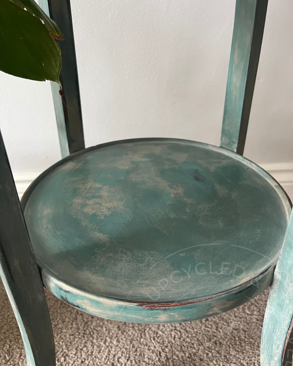The circular shelf of a two level distressed teal-green plant stand come side table. The upcycled plant stand has a soft, aged appearance with layers of greens, blues and cloudy whites showing
